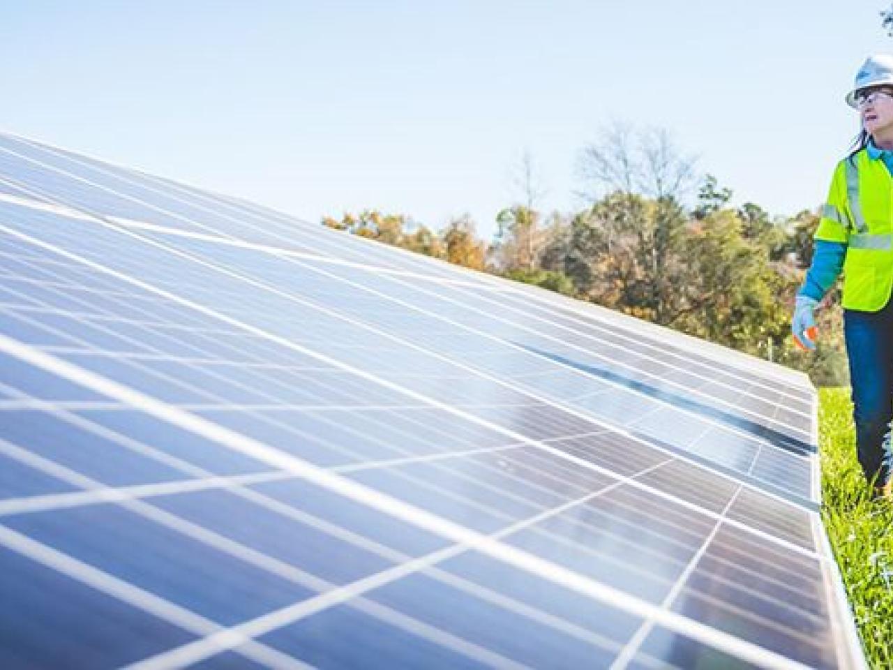 A person in a high-vis vest and hard hat inspects a solar panel on the ground.