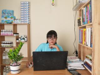 Thao sitting at a desk with a laptop. Shelves of books and products around her.