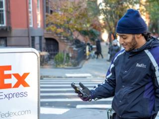 一个穿着FedEx制服的人旁边有FedEx Express徽标的高高推车。他们正在寻找住宅区的人行道上的手持设备。