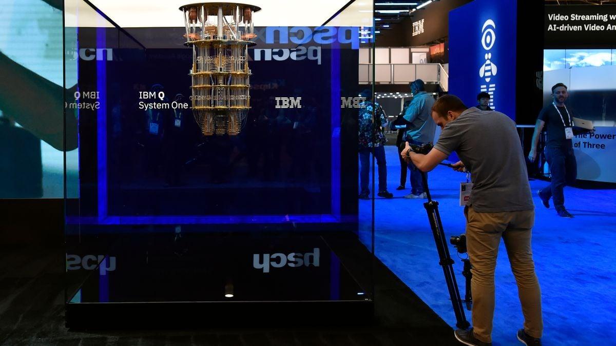 A person using a camera on a tripod to photograph a large device hanging from the ceiling of an glass enclosed space. 