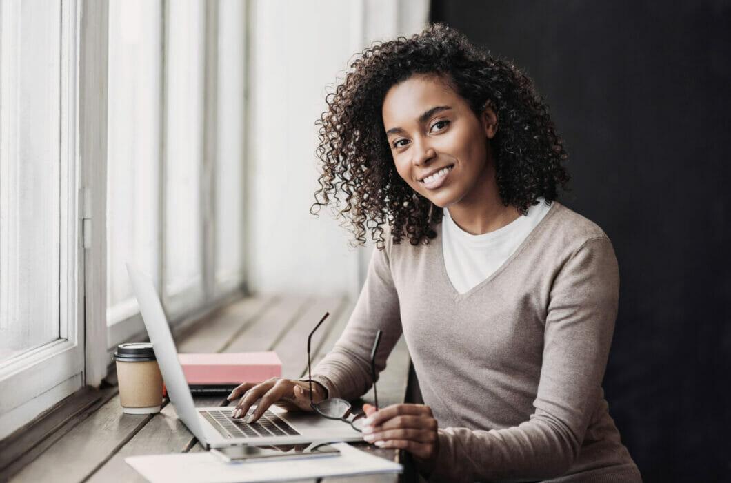 Young woman sitting at a desk, looking at the camera and typing on her laptop.