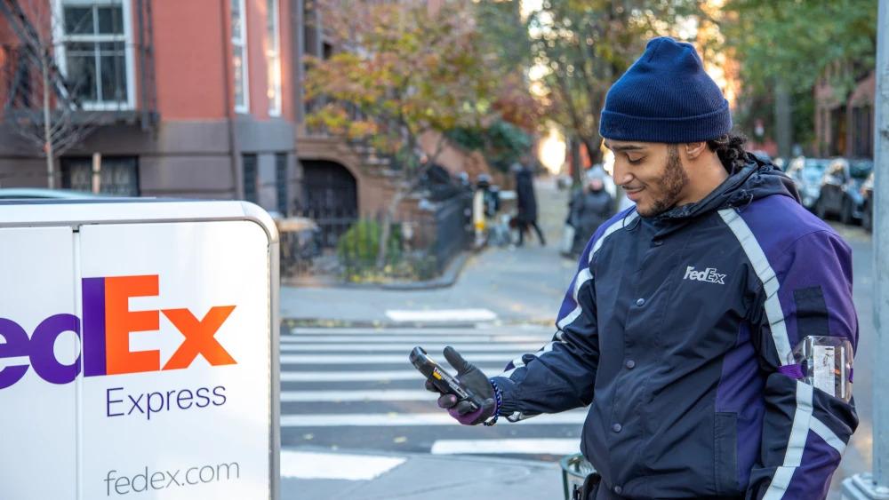 一个穿着FedEx制服的人旁边有FedEx Express徽标的高高推车。他们正在寻找住宅区的人行道上的手持设备。