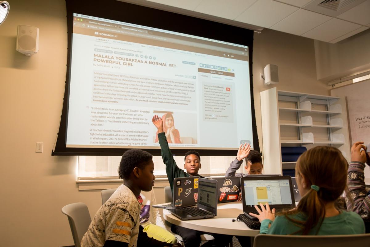 students at a round table, each on a laptop. A large pull-down projection screen behind them with a presentation on it.